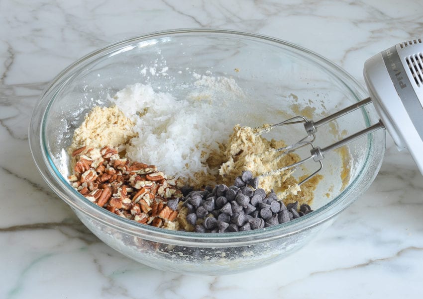 Chocolate chips, coconut, and nuts in a bowl with cookie dough.
