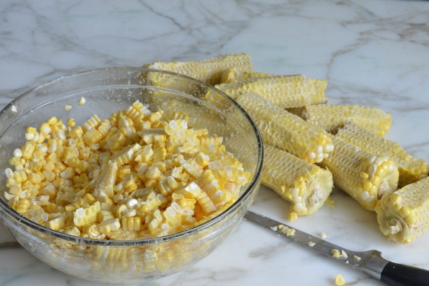 Bowl of corn kernels next to corn cobs.