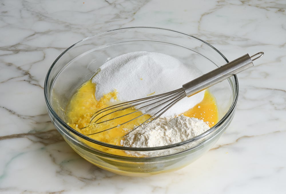 Adding sugar, lemon, and flour to eggs for filling in a bowl with a whisk resting on the side.