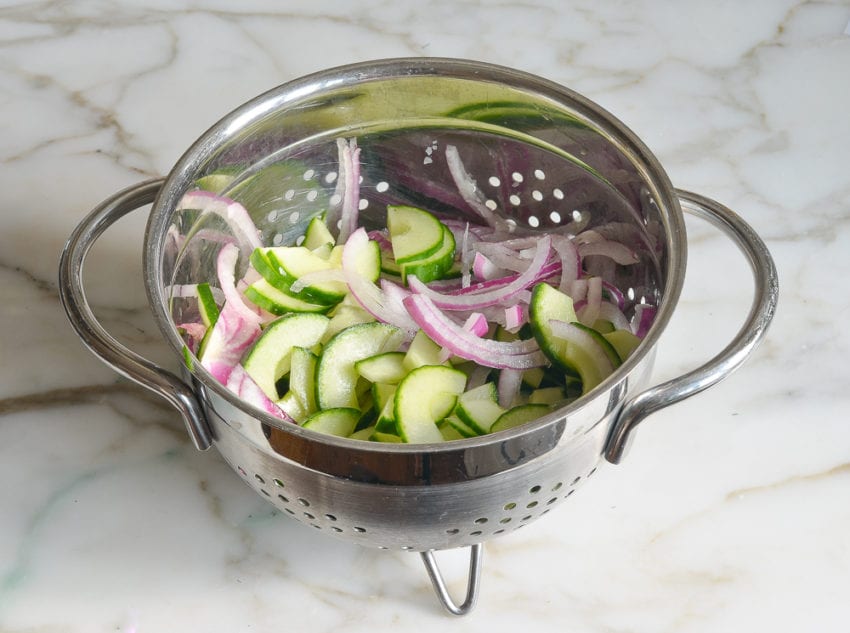 Sliced cucumbers and red onion in a colander.