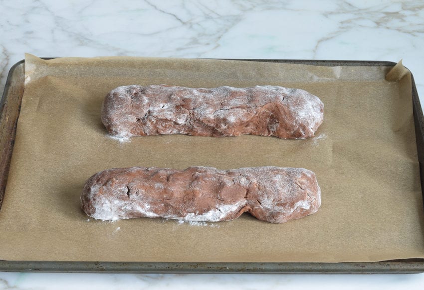 Two short logs of chocolate biscotti dough on a lined baking sheet.