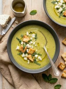 Bowls of pea and asparagus soup on wooden board with feta, mint, and pita croutons.