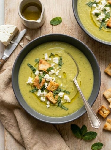 Bowls of pea and asparagus soup on wooden board with feta, mint, and pita croutons.