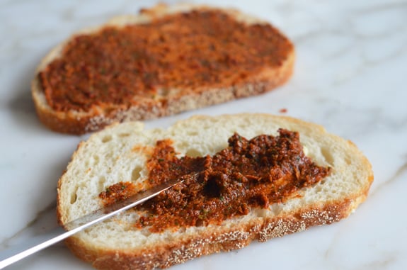Knife spreading sun-dried tomato pesto on bread.