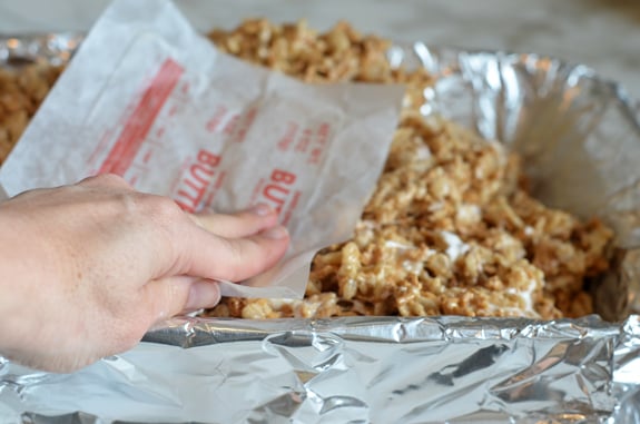 Person pressing cereal mixture into a lined baking pan with a butter wrapper.