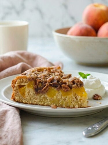 Peach cake on plate with bowl of peaches and coffee mug in background.
