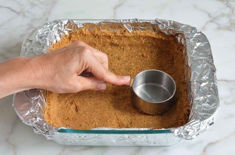 Person pressing graham cracker crust mixture into a lined baking pan with a measuring cup.