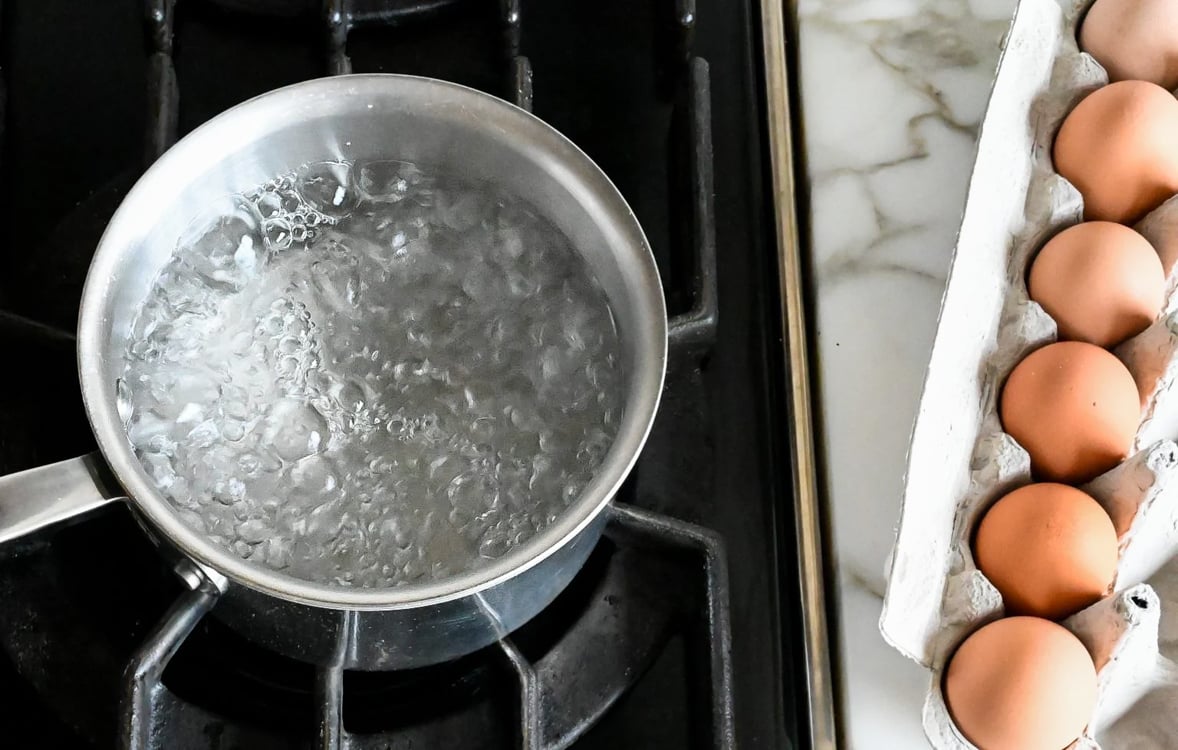 Boiling water on stove with carton of eggs on counter.