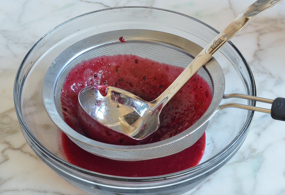 Spoon pushing a berry mixture through a sieve.