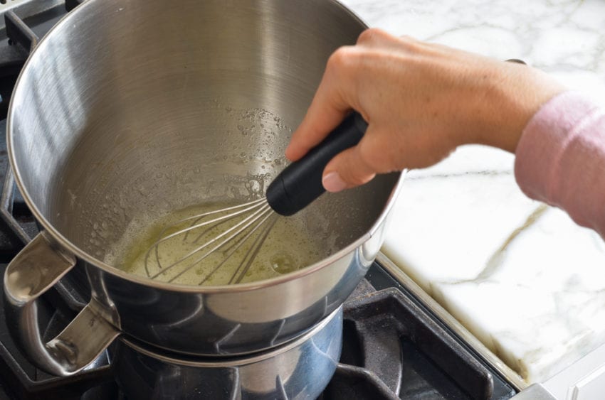 Person whisking egg whites and sugar in a bowl on a saucepan on a stovetop.