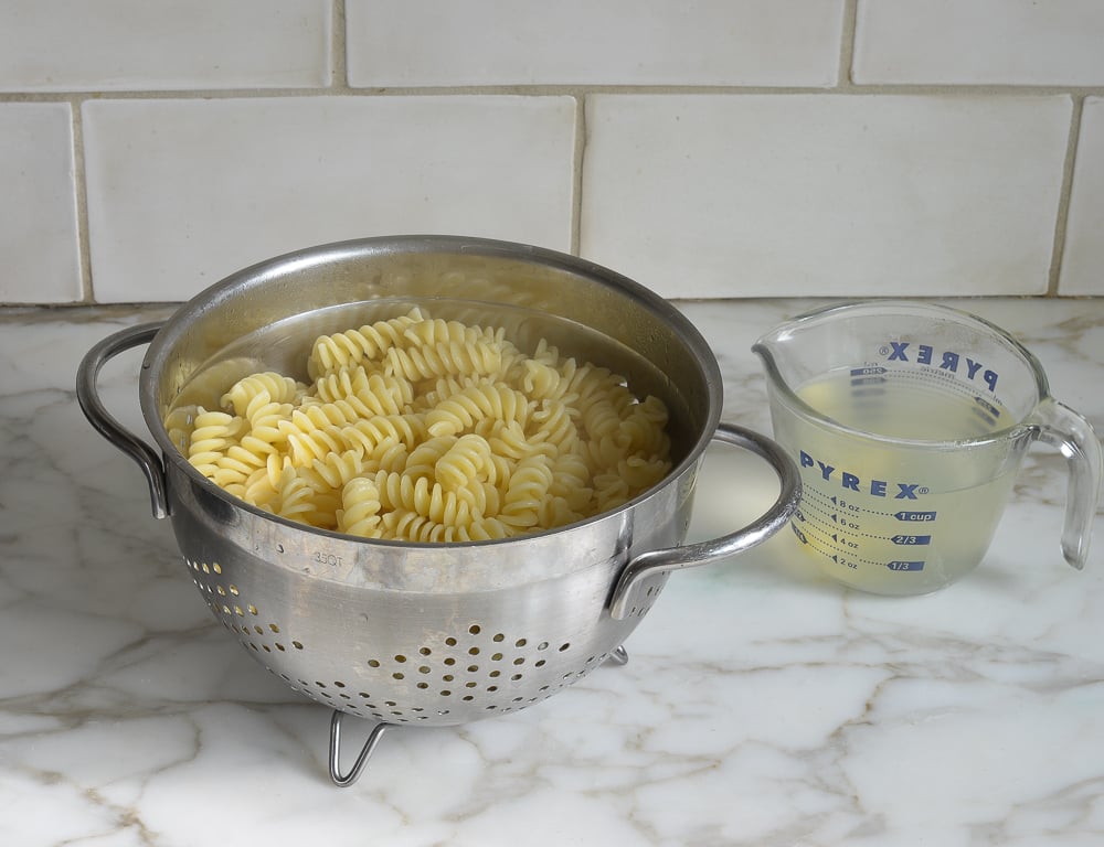 Colander of pasta next to a measuring cup of pasta water.
