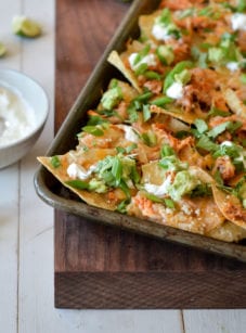Sheet pan chicken nachos on a wooden board with a bowl of sauce and lime wedges to the side.