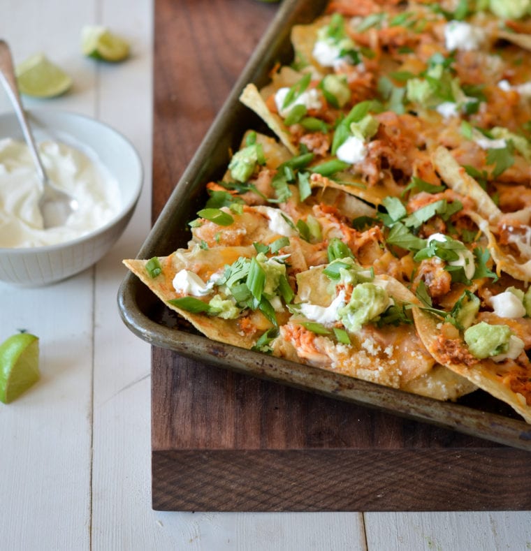 Sheet pan chicken nachos on a wooden board with a bowl of sauce and lime wedges to the side.