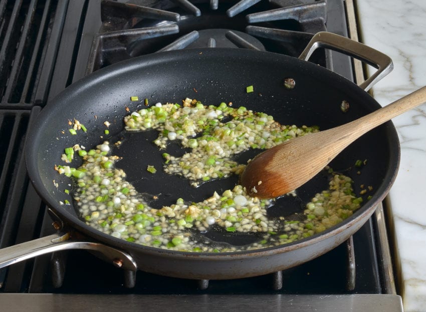 Spoon stirring scallions, ginger, and garlic in a skillet.