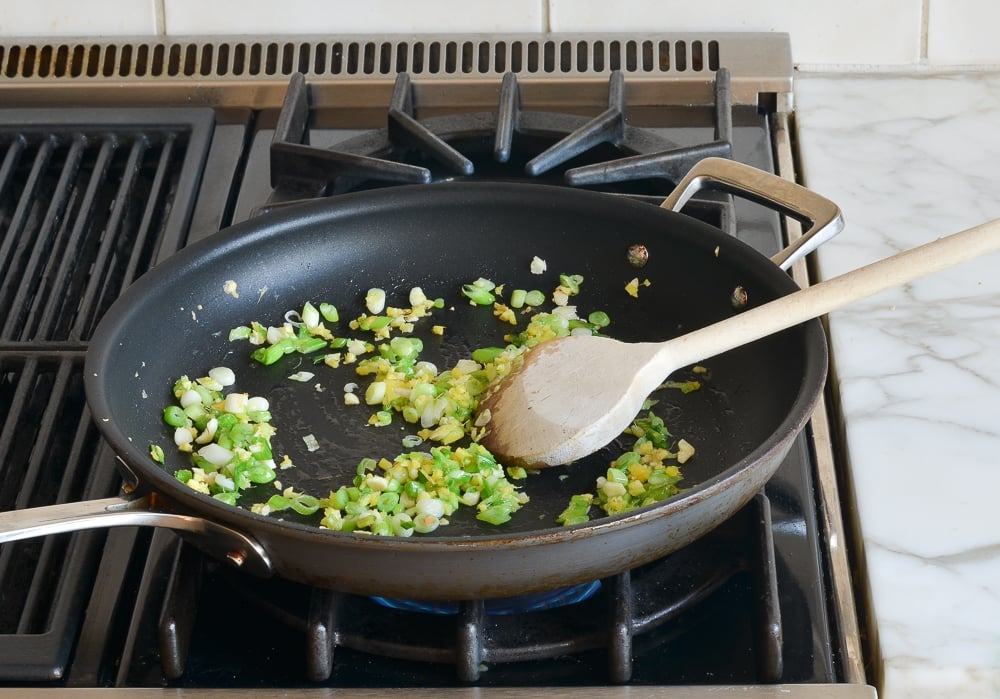 Spoon stirring scallions, garlic, and ginger in a skillet.