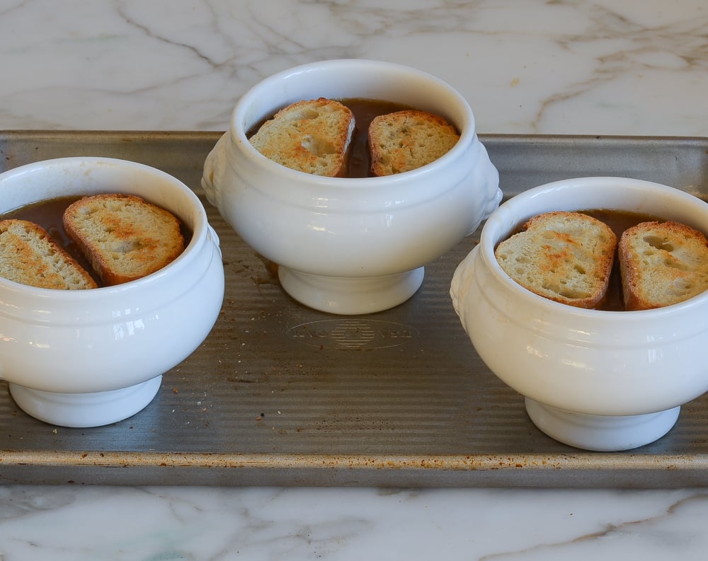 French onion soup crocks with baguette slices on baking sheet.