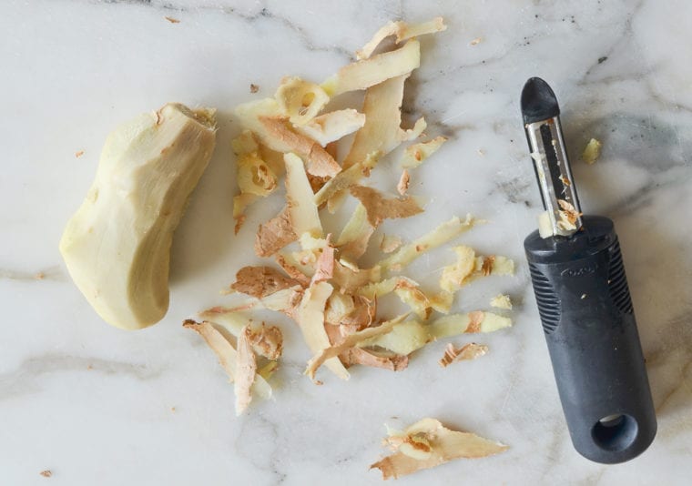 Vegetable peeler next to a pile of ginger peels and a peeled ginger root.