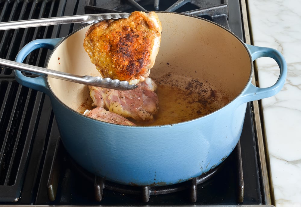 Tongs flipping browned chicken in a Dutch oven.