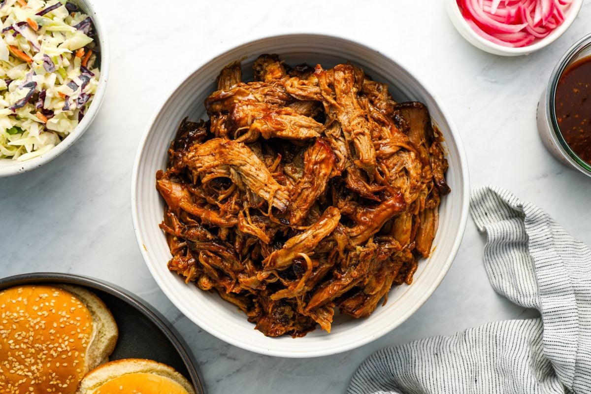 pulled pork in bowl surrounded by sandwich buns, coleslaw and pickled onions