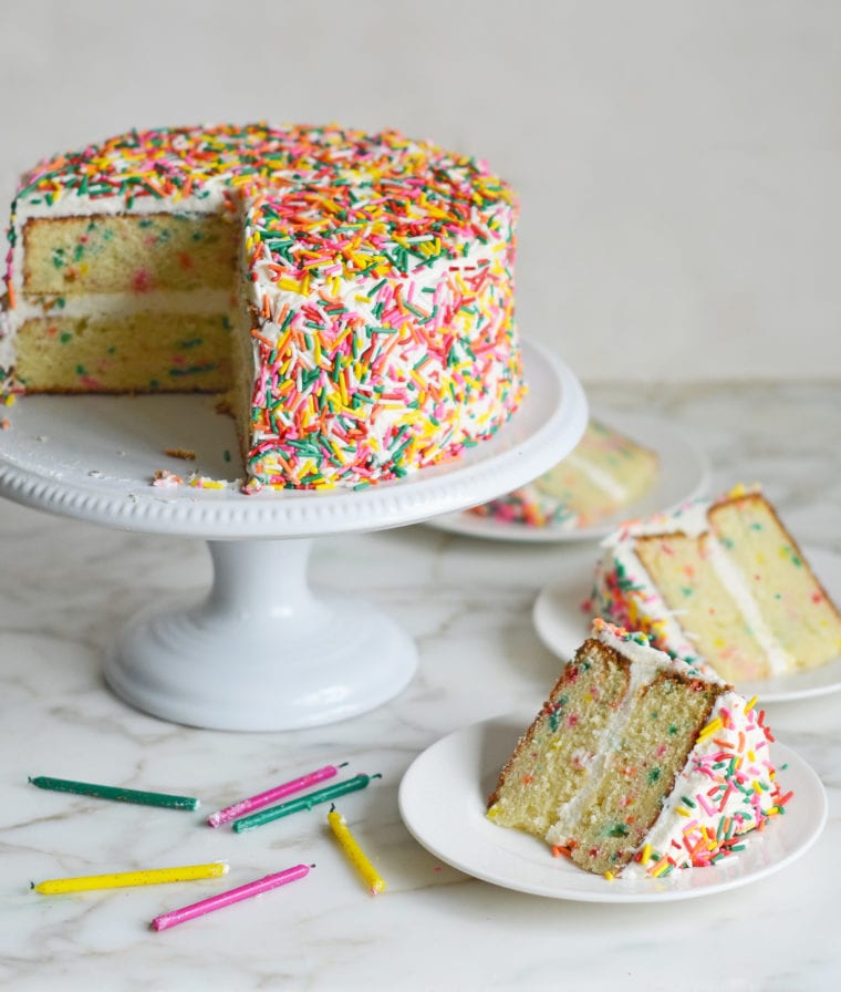 Slices of rainbow sprinkle Funfetti cake on plates next to the rest of the cake.