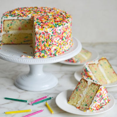 Slices of rainbow sprinkle Funfetti cake on plates next to the rest of the cake.