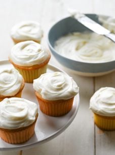 Vanilla cupcakes on and around a raised platter.