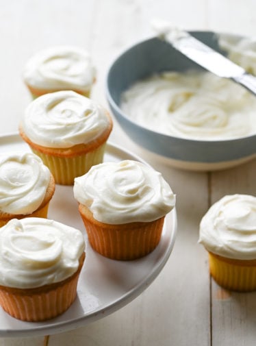 Vanilla cupcakes on and around a raised platter.