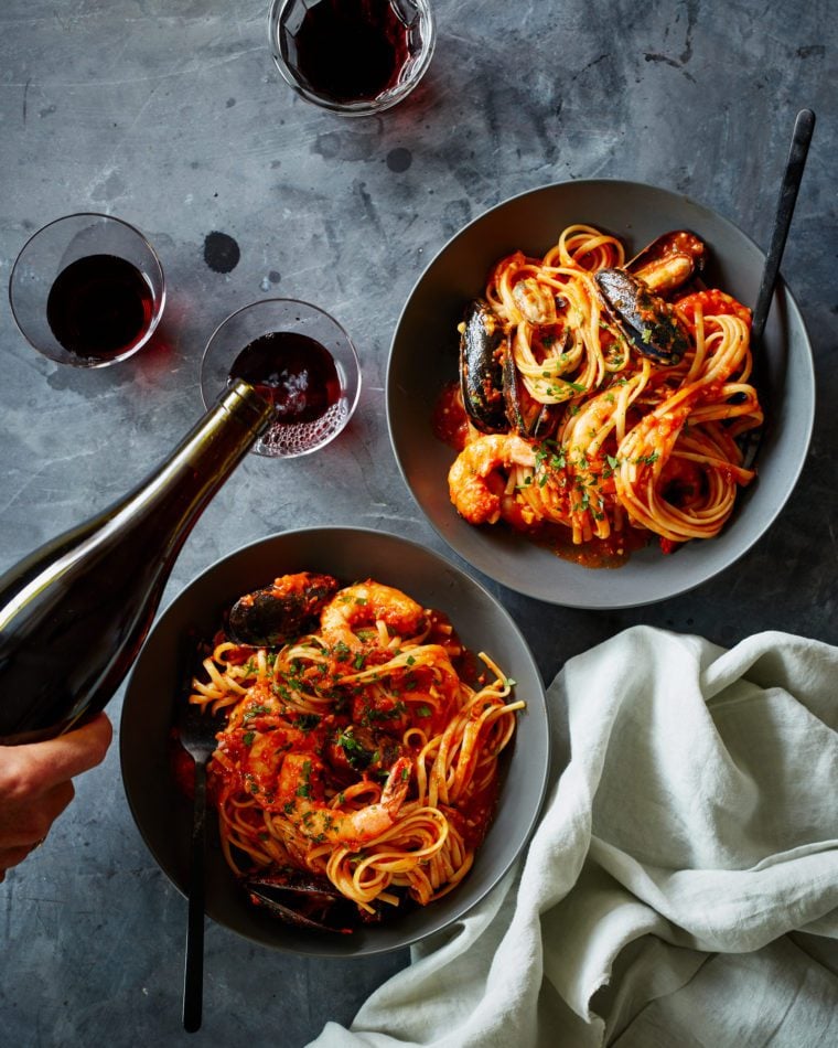 Bottle pouring red wine into a glass with plates of pasta.