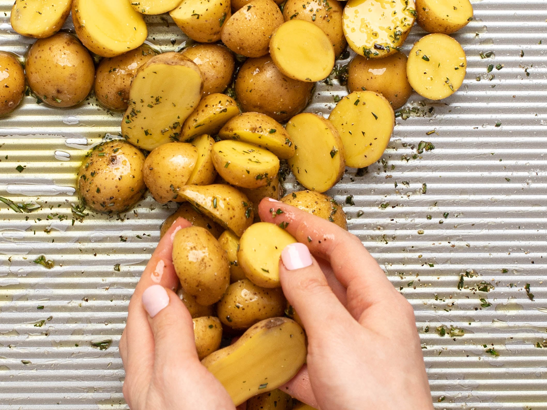 Hands tossing the potatoes with oil and seasonings on a baking tray.
