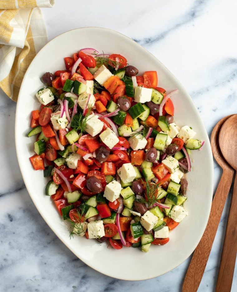 A large platter of Greek salad on a table with a wooden spoon to the side.