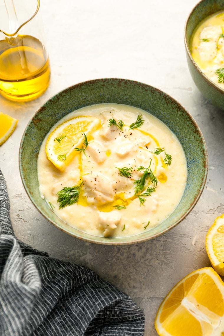 Avgolemono soup in bowl with gray striped dish towel, olive oil, and lemon wedges.