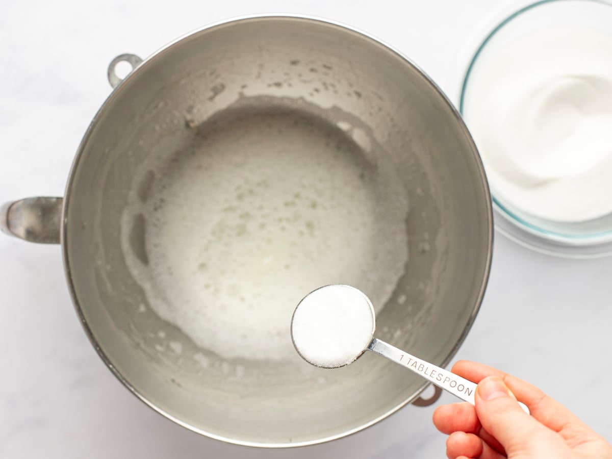 Adding sugar to beaten egg whites, salt, and cream of tartar in mixing bowl.