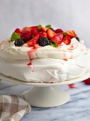 Pavlova topped with macerated berries on a cake stand with a linen napkin to the side.