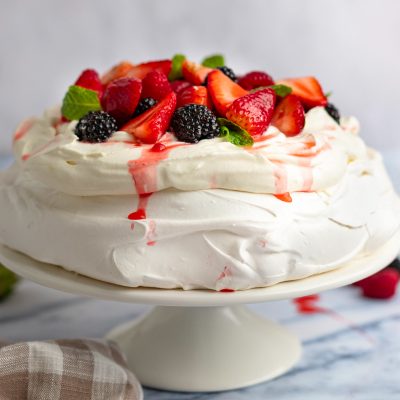 Pavlova topped with macerated berries on a cake stand with a linen napkin to the side.
