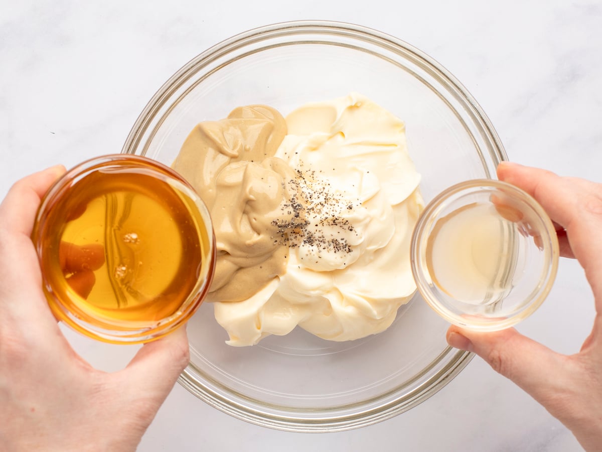 pouring vinegar and honey into glass bowl containing mustard, mayonnaise, and black pepper