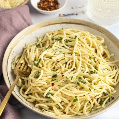spaghetti aglio e olio in serving bowl with fork; bowl has a mauve napkin to the left and small bowl with red pepper flakes above