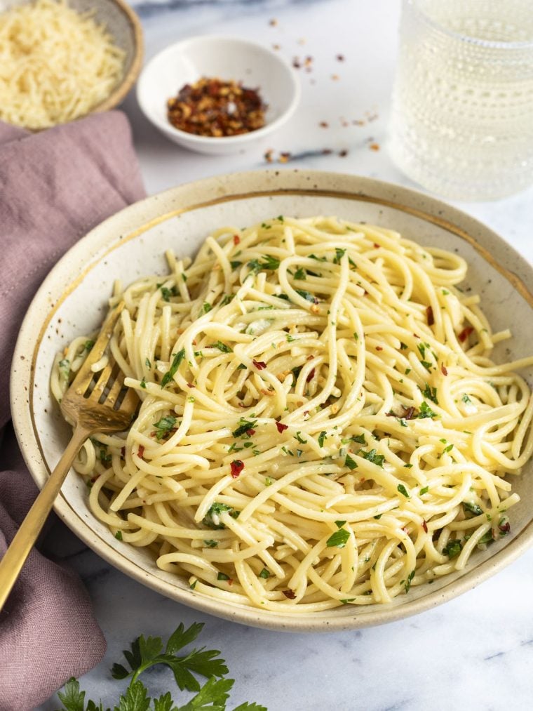spaghetti aglio e olio in serving bowl with fork; bowl has a mauve napkin to the left and small bowl with red pepper flakes above