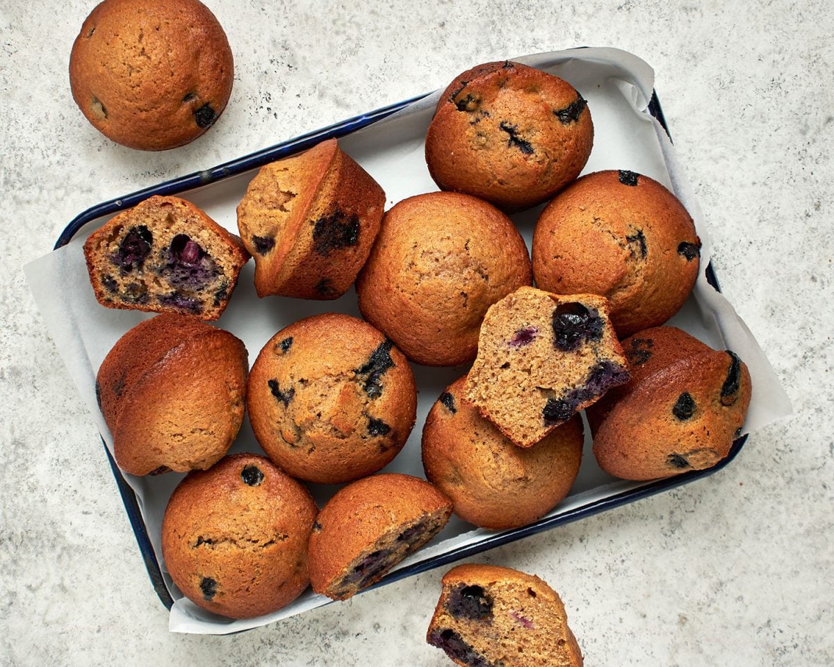 blueberry maple bran muffins piled onto baking sheet with additional muffins on the counter nearby