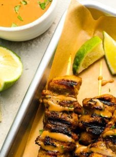 small white bowl with thai curry peanut sauce next to baking sheet with chicken skewers drizzled with the sauce