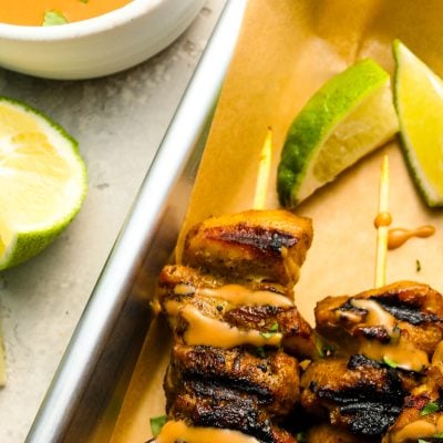 small white bowl with thai curry peanut sauce next to baking sheet with chicken skewers drizzled with the sauce