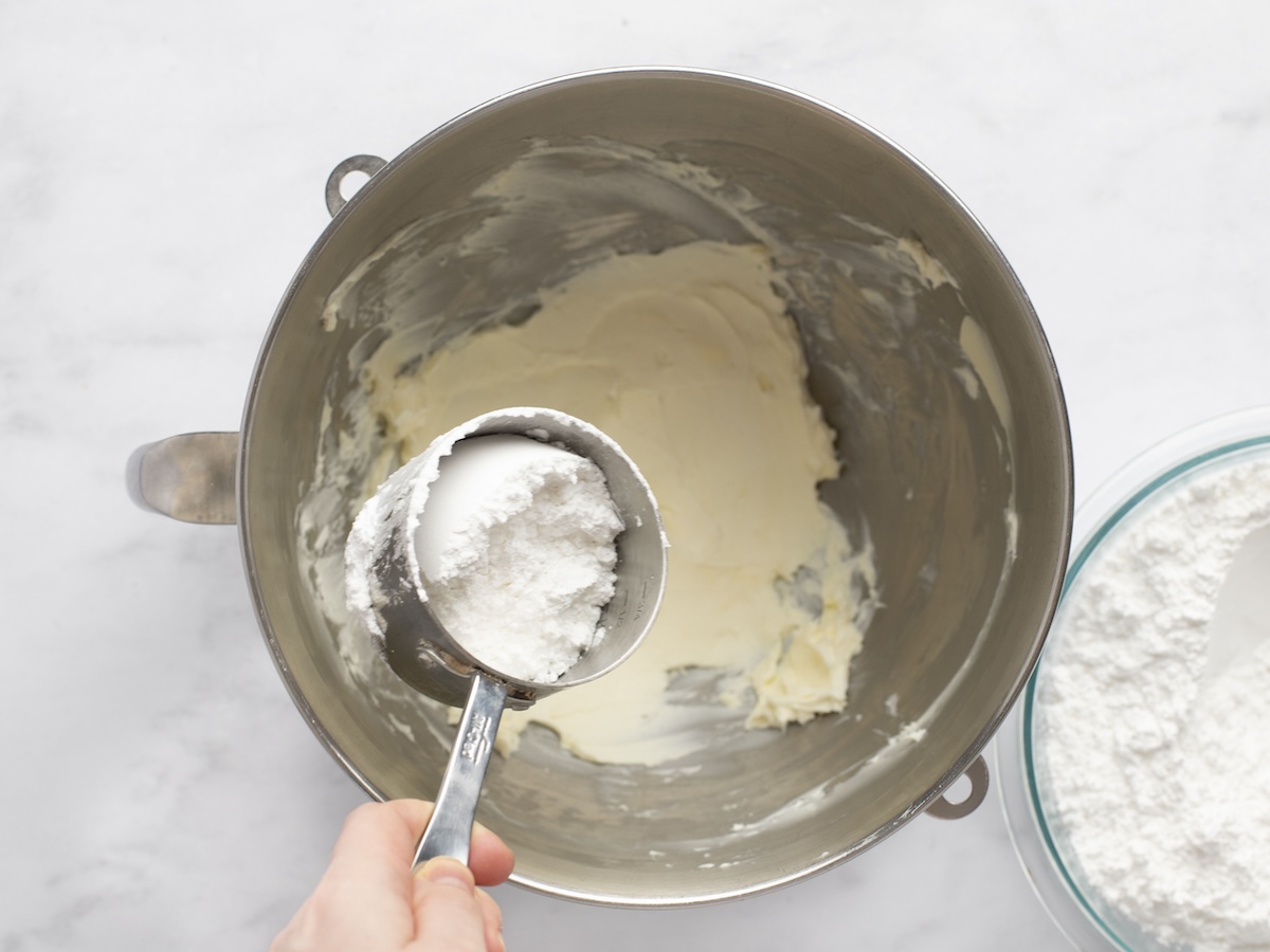 mixing bowl with confectioners' sugar being added to beaten butter and cream cheese