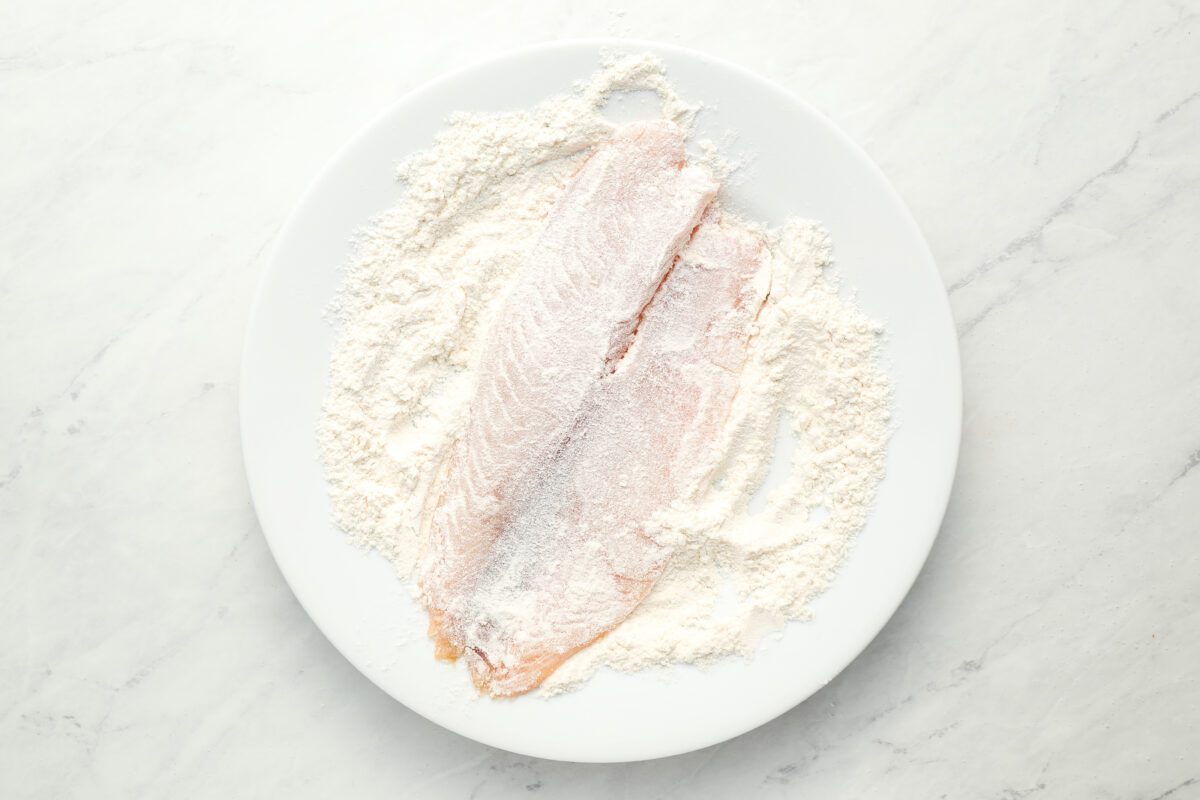 dredging 1 tilapia fillet in flour in white bowl