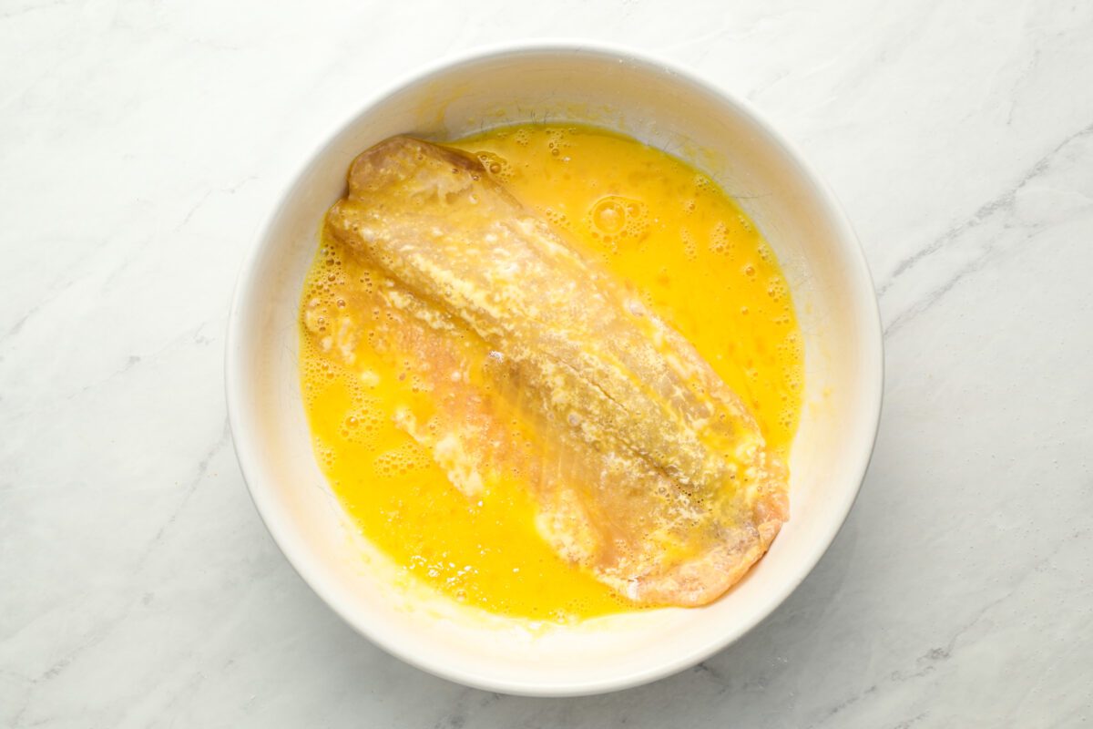 dipping flour covered fish in beaten eggs in white bowl