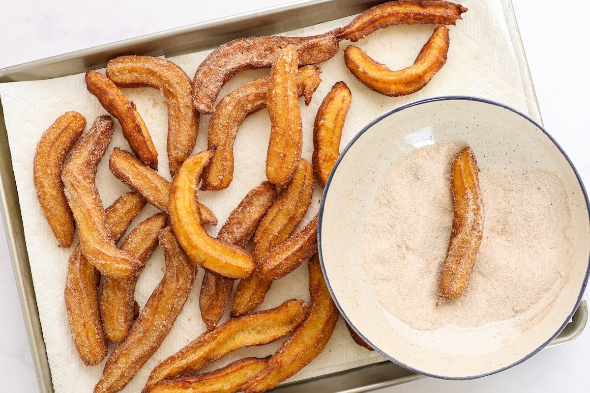 bowl of cinnamon sugar and finished churros covered in cinnamon sugar on paper towel-lined baking sheet