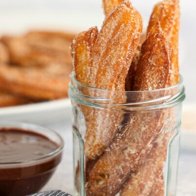 churros in glass jar with chocolate sauce and platter of churros in background.