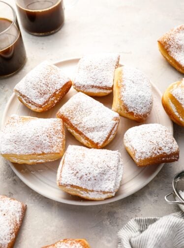 sugar-dusted beignets on round white plate with 2 cups of coffee