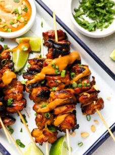 white serving platter with cooked chicken skewers and small bowl of peanut sauce