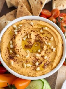 hummus topped with pine nuts, sumac, and olive oil in white serving bowl; bowl is placed between fresh vegetables and pita triangles