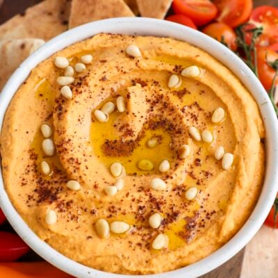 hummus topped with pine nuts, sumac, and olive oil in white serving bowl; bowl is placed between fresh vegetables and pita triangles