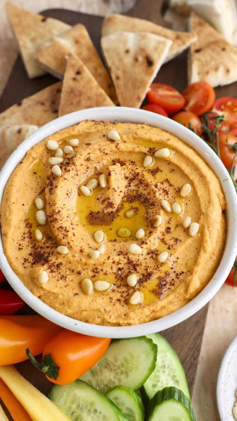 hummus topped with pine nuts, sumac, and olive oil in white serving bowl; bowl is placed between fresh vegetables and pita triangles